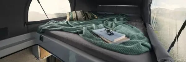 Gemachtes Bett im Schlafbereich eines Camper Vans mit grüner Decke, Kissen, Buch und Brille. Ausblick auf Berge durch geöffnete Seitenfenster.