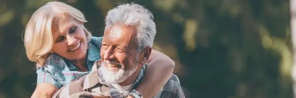 Ein altes Ehepaar im Grünen. Er nimmt sie huckepack. Beide lächeln. Die Sonne scheint.