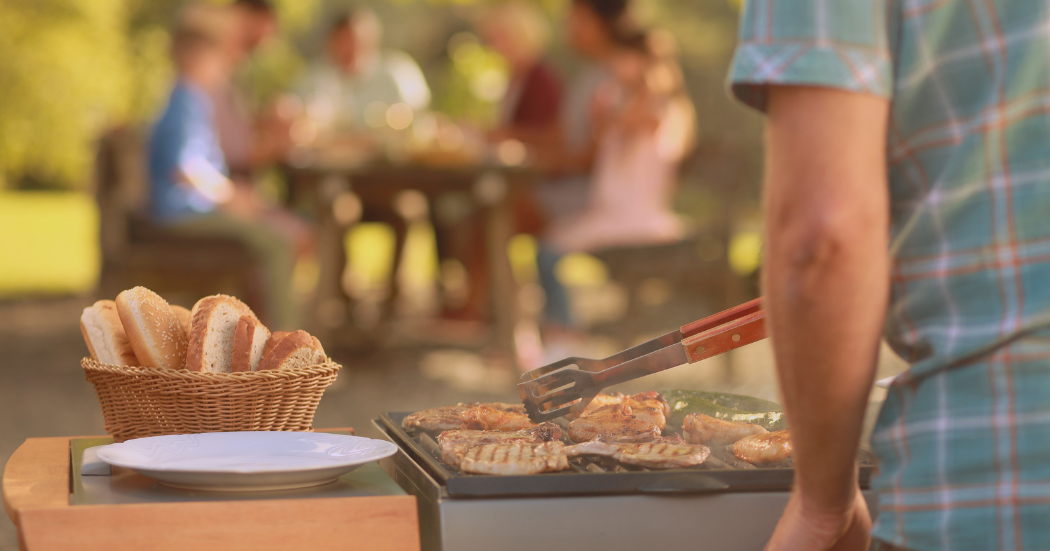 Ein Mensch steht am Grill. Er wendet das Grillgut. Im Hintergrund sitzen gesellige Menschen und essen das zubereitete Grillgut. Ein Mensch steht am Grill. Er wendet das Grillgut. Im Hintergrund sitzen gesellige Menschen und essen das zubereitete Grillgut.